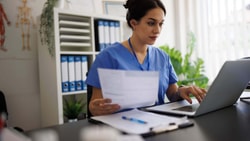 Outpatient clinician reviewing remote therapeutic monitoring (RTM) denied claims and billing documents on laptop in clinic office