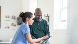 Clinician in blue scrubs showing an older male patient RTM data on a tablet in an outpatient clinic.