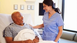 A female nurse smiling and supporting an elderly male patient in a hospital bed; reducing hospital readmissions.