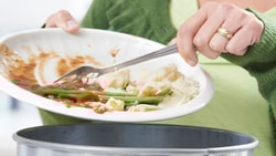 Person in a green sweater scraping food scraps from a plate into a trash can.