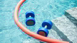 Aquatic therapy equipment in a pool, including foam dumbbells and a noodle, used to support pediatric exercise and conditioning.