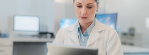 A woman in a white lab coat uses a tablet in a modern medical office, representing digital healthcare and medbridge trends.