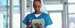 A healthcare worker in blue scrubs uses medbridge software on a tablet while standing indoors.