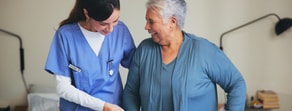 A nurse helps a smiling older woman stand in a bright room with medical gear and digital medbridge tools in the background.
