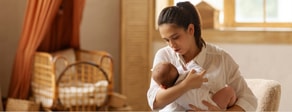 A woman in a white shirt breastfeeds her baby indoors, with a crib and window behind them, illustrating medbridge healthcare.