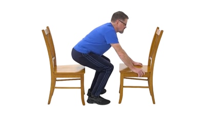 A man uses two wooden chairs to perform a sit-to-stand exercise for medbridge healthcare training.