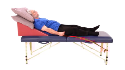 An older man reclines on a therapy table with a red resistance band on his ankle during medbridge training.