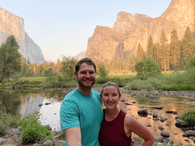Two people smiling for a selfie by a river with cliffs and trees, under clear sky, representing medbridge hybrid care.