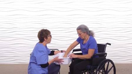 A caregiver assists a woman in a wheelchair with hand soaking, highlighting hybrid care, against a wavy patterned wall.