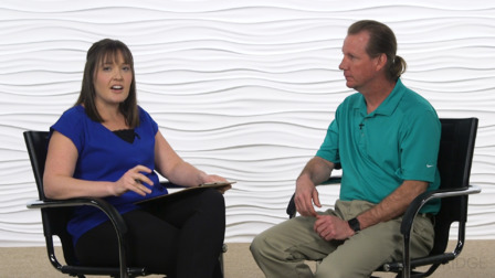 A woman and man discuss healthcare education seated on black chairs by a white textured wall.