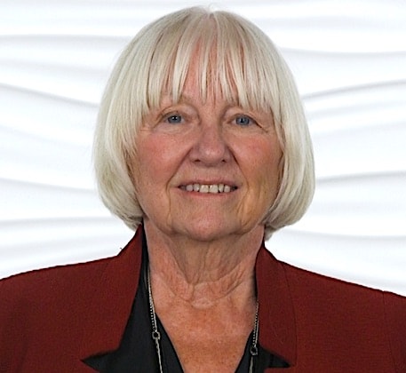 An elderly person with short white hair and a red jacket smiles in front of a wavy white background.