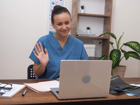 Clinician conducting remote therapeutic monitoring (RTM) telehealth visit, waving during video call on laptop in clinic office
