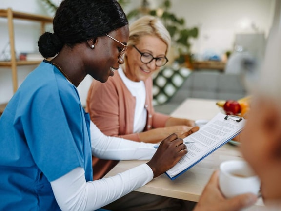 A healthcare worker in scrubs talks with an older woman about digital healthcare solutions at a table, possibly using medbridge.