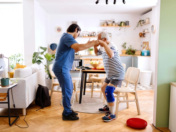 Male OT helping an elderly man practice a sit-to-stand transition in a home kitchen setting.