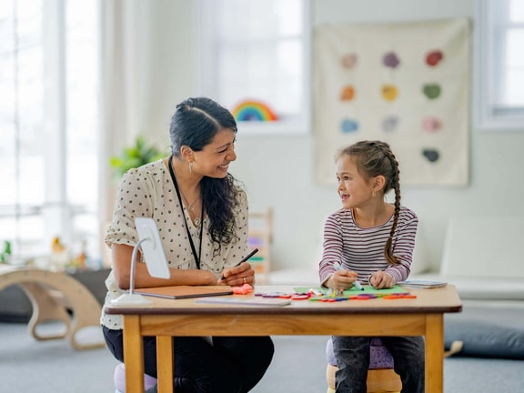 A smiling SLP and young girl engage in a play-based autism assessment at a small wooden table.