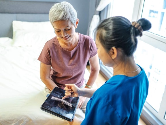 A healthcare worker shows patient info on a tablet to an older woman sitting on a bed; screen displays medbridge software.