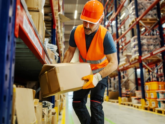 Worker in orange safety vest and helmet lifts a box in warehouse supporting medbridge healthcare training and hybrid care.