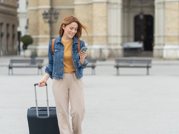 Travel therapist with suitcase using a phone to manage licensure and CEU requirements in a city plaza.