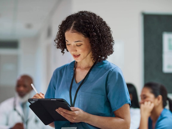 Nurse reviewing a tablet while completing a nursing skills checklist in a clinical setting.