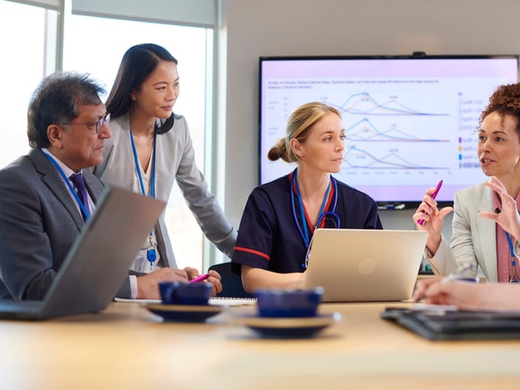 Healthcare professionals in a meeting discussing PEPM vs PMPM strategies using data charts on a screen.