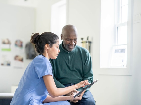 Clinician in blue scrubs showing an older male patient RTM data on a tablet in an outpatient clinic.