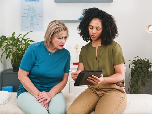 A healthcare worker shows medbridge info on a tablet to an older woman in a medical office.