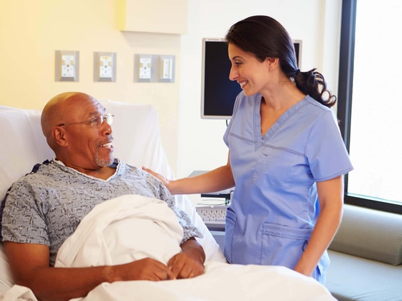 A female nurse smiling and supporting an elderly male patient in a hospital bed; reducing hospital readmissions.