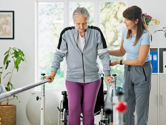 Therapist assisting a senior patient with gait training in parallel bars to address Unsteadiness on Feet (R26.81).
