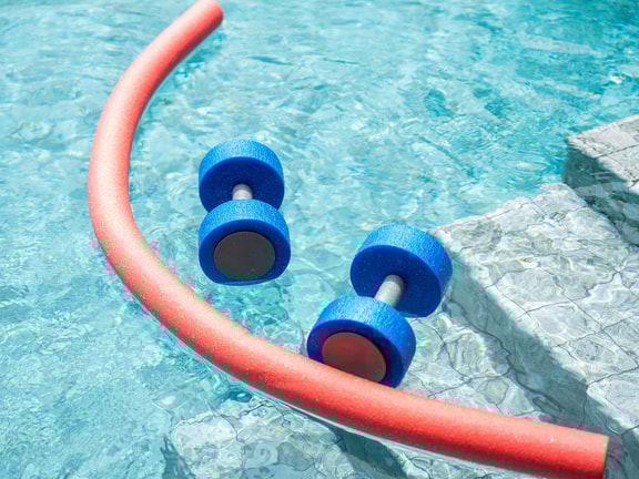 Aquatic therapy equipment in a pool, including foam dumbbells and a noodle, used to support pediatric exercise and conditioning.