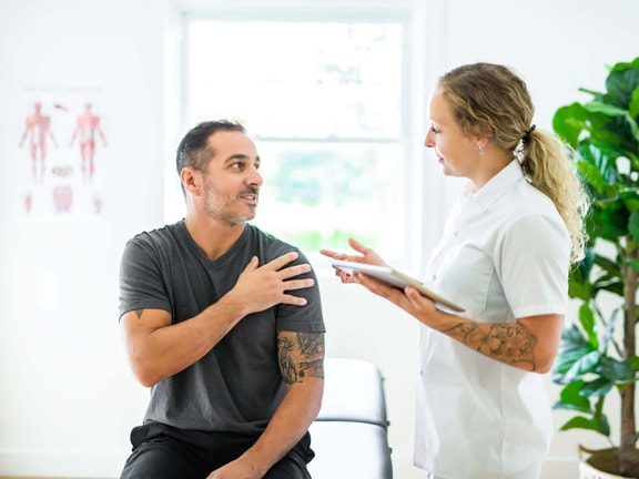A female clinician discusses chronic shoulder pain with a male patient, applying a functional medicine framework.