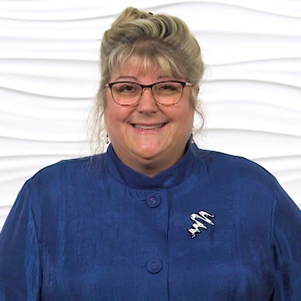 A person with glasses and a blue jacket smiles against a white textured wall for medbridge.