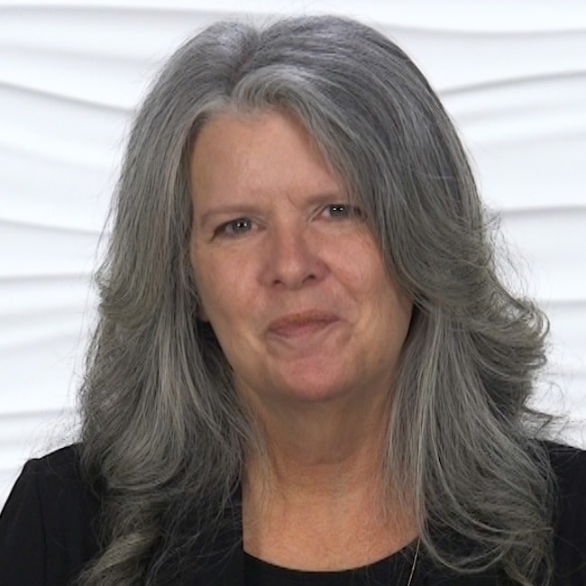 A woman with long gray hair smiles slightly in front of a textured white background, wearing a black top.