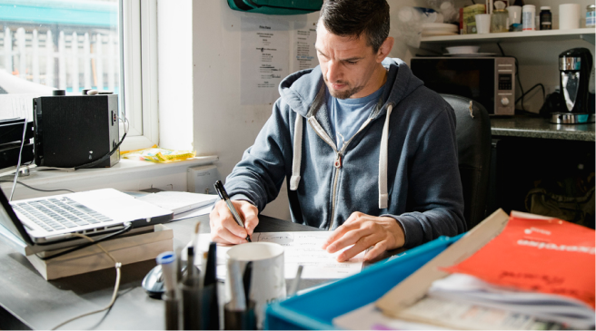 Man in a hoodie works at a messy desk with papers and laptop, showing digital healthcare and hybrid care at medbridge.