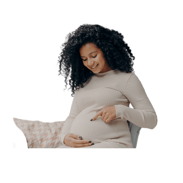 Smiling pregnant woman with curly hair, pointing to her belly and a pillow in the background.