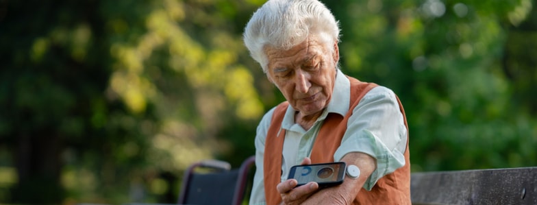 An older adult scans a glucose monitor on their arm with a device while sitting on an outdoor bench.
