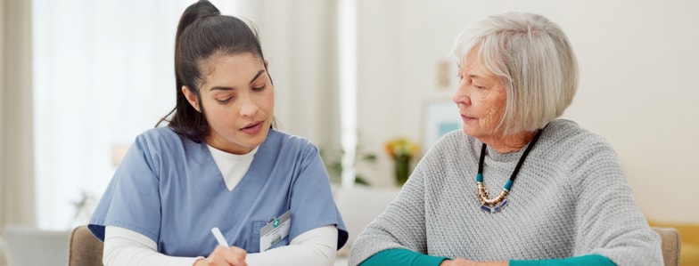 A healthcare worker in scrubs writes on a clipboard next to an older woman, showing attentive support with medbridge hybrid care.