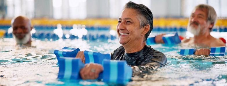 Three older adults use foam dumbbells in a pool during water aerobics, promoting wellness and medbridge digital health.