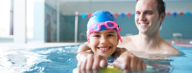 Child in swim cap and goggles holds a kickboard in a pool next to an adult, showing medbridge water safety education.