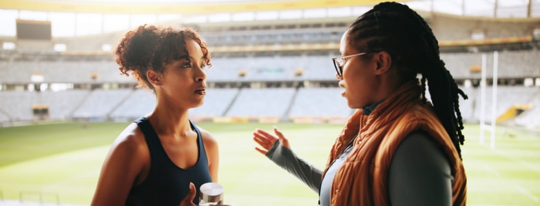 Two women talk at a stadium near an empty rugby field, discussing digital healthcare and medbridge.