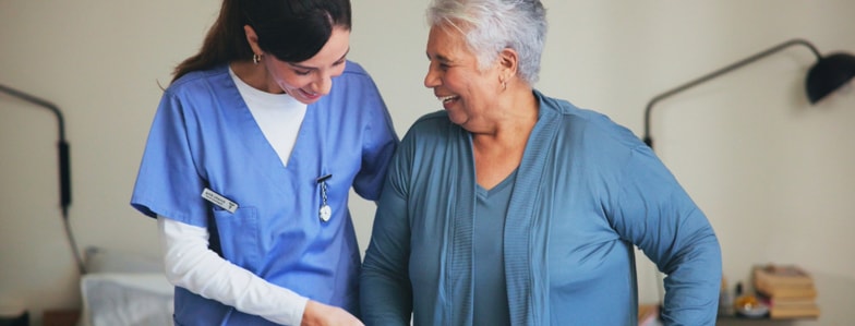 A nurse helps a smiling older woman stand in a bright room with medical gear and digital medbridge tools in the background.