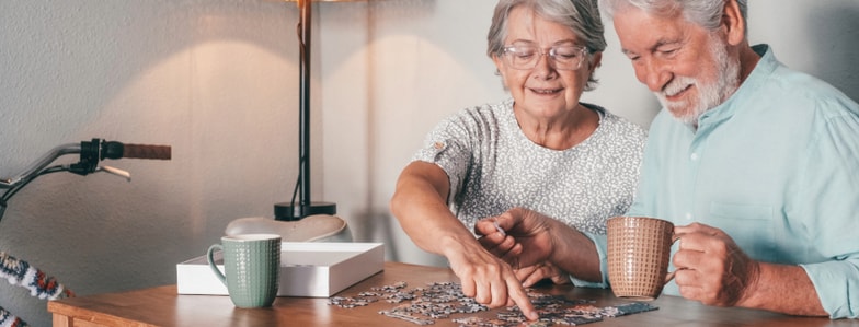 An elderly couple assembles a puzzle at a table with mugs, a lamp, and bike nearby, suggesting everyday medbridge healthcare.
