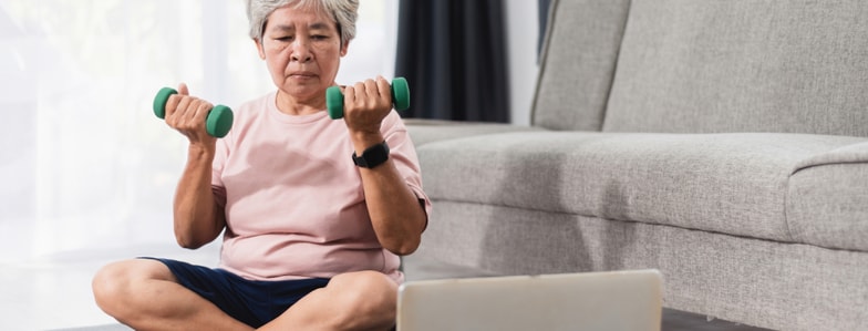 Older adult lifts dumbbells while following medbridge training on a laptop, seated on the floor near a gray sofa.