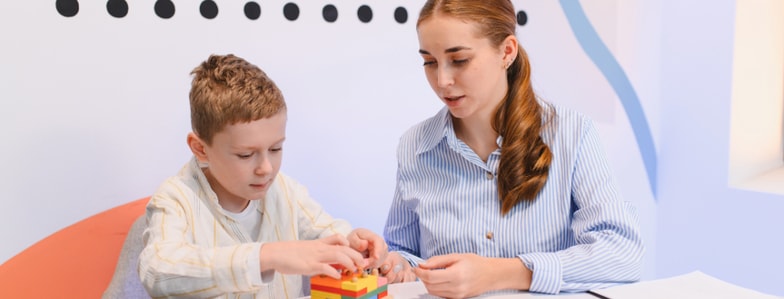 A woman helps a young boy stack blocks at a table with papers, using medbridge software for his learning.