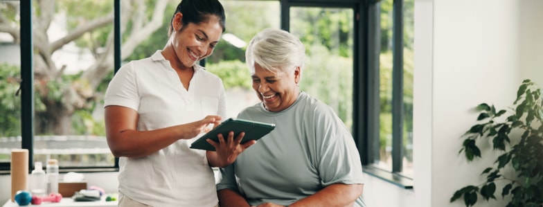 A younger woman helps an older woman use a tablet in a bright room, showing the benefits of digital healthcare with medbridge.