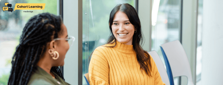 Two women smile at each other indoors near a "Cohort Learning" sign, emphasizing medbridge digital healthcare training.