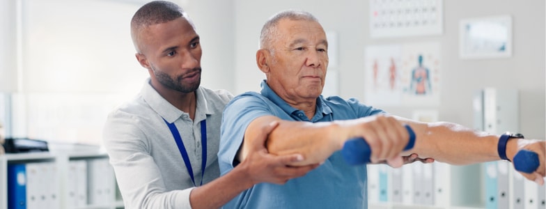 A physical therapist uses medbridge software to help an older man lift light dumbbells during rehab in a clinic.