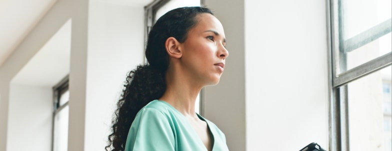 A woman with curly hair in a green top looks thoughtfully out a window, reflecting on her medbridge healthcare training.