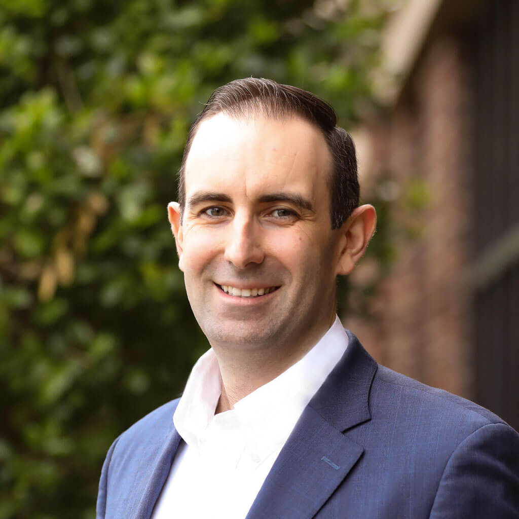 A man in a blue suit smiles outdoors against a backdrop of green foliage, envisioning the future of digital healthcare with Medbridge.