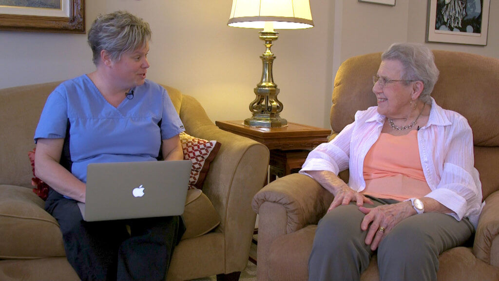 A healthcare worker in blue scrubs talks to an older woman holding a laptop, promoting digital healthcare with medbridge in a warm setting.