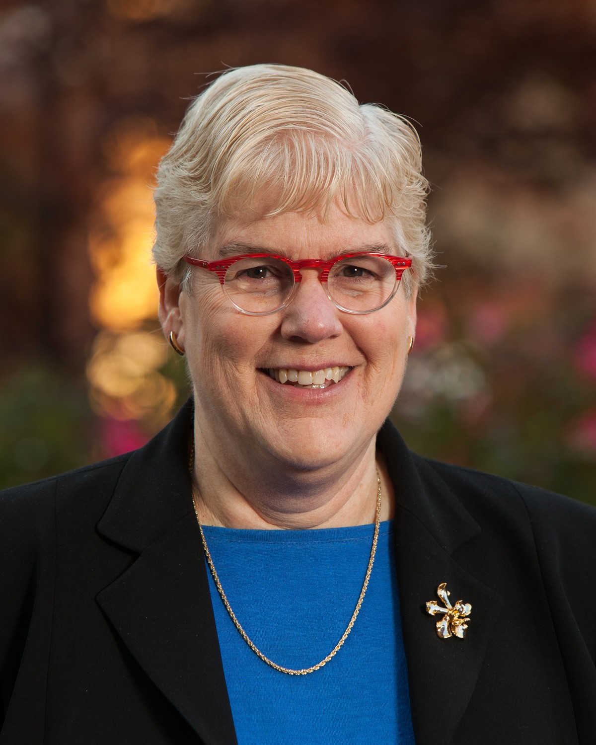 A smiling person with short white hair, glasses, and a black blazer with a floral pin is outdoors.